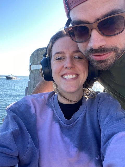 In this image, Lisa is smiling, wearing headphones and a purple sweatshirt. Another person is next to her, wearing sunglasses and a cap. They appear to be outdoors near a body of water, with a boat visible in the background.
