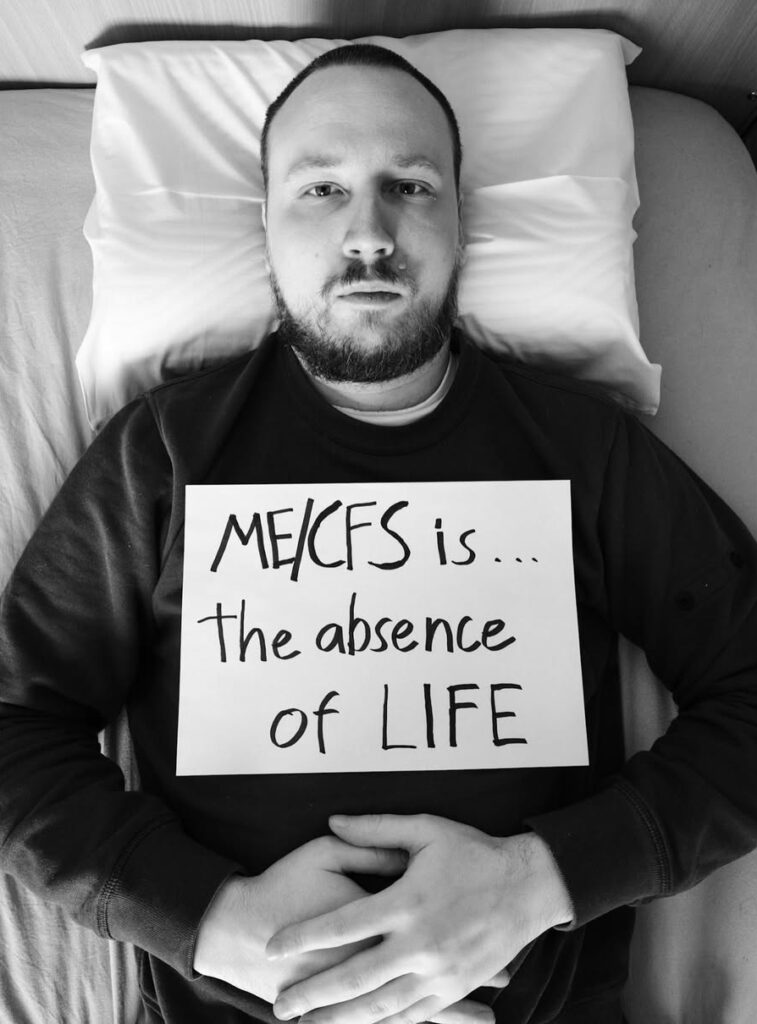 Niko is holding a sign that reads: ME/CFS is the absence of life.