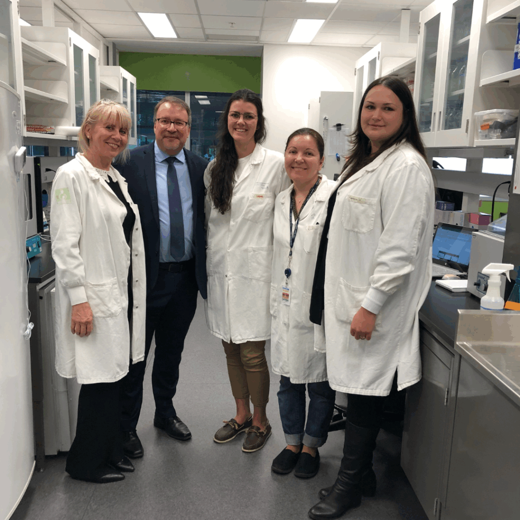 The image shows Dr. Alain Moreau and four members of his research team at OMF’s Collaborative Center in Montreal. They are standing together in a laboratory setting. Dr. Moreau is dressed in a suit, while the rest of the team are wearing white lab coats.