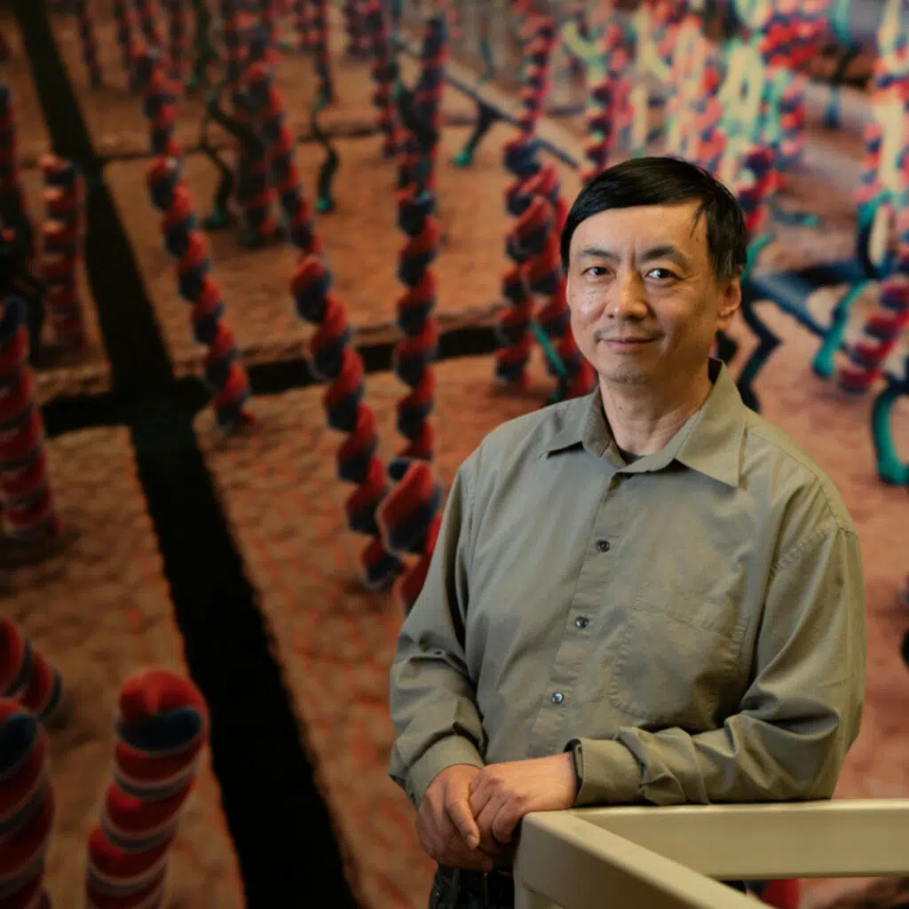 The image shows Wenzhong Xiao standing indoors, leaning lightly on a railing. Behind him is a large, colorful scientific display featuring oversized, twisted, rope-like structures in red and blue, arranged in repeating rows.